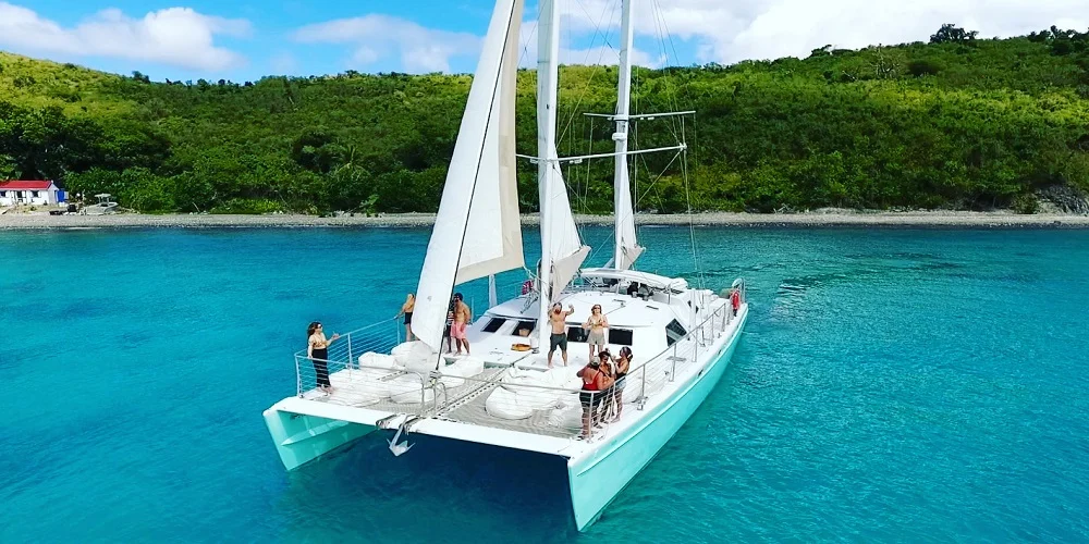 Catamaran sailing coast with guests near lush shoreline.